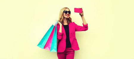 Portrait of beautiful smiling young woman taking selfie with smartphone holding shopping bags on...