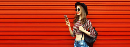Portrait of happy smiling young woman with smartphone wearing black round hat on red background