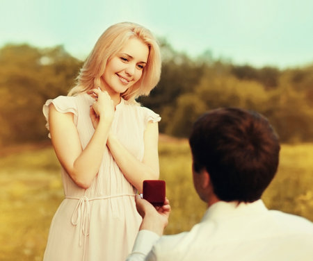 Happy young couple, man kneeling down and proposing a ring to his beloved woman outdoors in the park, wedding conceptの写真素材