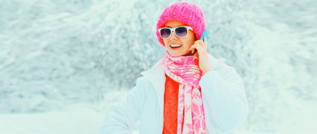 Winter portrait of happy smiling young woman calling on smartphone looking away wearing knitted hat and jacket in the park on snowy backgroundの写真素材