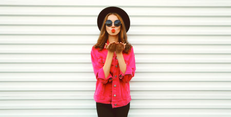Portrait of stylish young woman model blowing her lips sending sweet air kiss wearing pink jacket, black round hat on white backgroundの写真素材