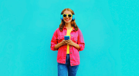 Portrait of stylish modern happy smiling young woman in headphones listening to music with smartphone wearing pink jacket on vivid blue backgroundの写真素材