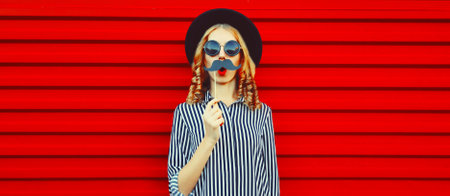 Portrait of funny cheerful young woman having fun showing mustache on stick on red backgroundの写真素材