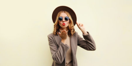Portrait of stylish beautiful young woman model blowing her lips sends kiss wearing brown round hat and coat on beige backgroundの写真素材
