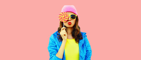 Portrait of stylish young woman blowing her lips with lollipop wearing colorful pink hat, blue jacket on pink backgroundの写真素材