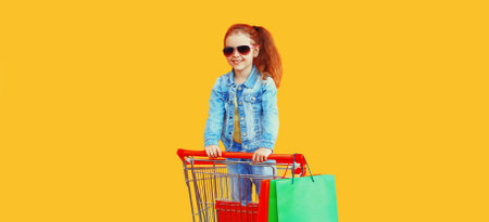 Portrait of happy smiling little girl child with trolley cart and shopping bags wearing denim clothes on yellow backgroundの写真素材