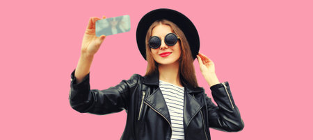 Portrait of beautiful smiling young woman taking selfie with smartphone posing wearing black round hat on pink backgroundの写真素材