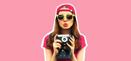 Portrait of happy young woman photographer taking picture on film camera and blowing her lips wearing baseball cap on pink background, magazine styleの写真素材
