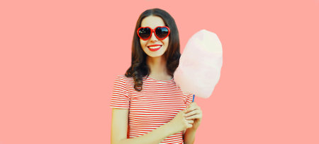Portrait of happy smiling young woman with cotton candy wearing red heart shaped sunglasses on pink...