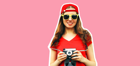 Portrait of happy smiling young woman photographer taking picture on film camera wearing red baseball cap on pink background, magazine styleの写真素材