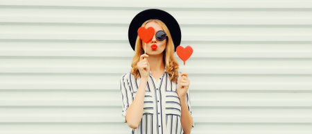 Portrait of beautiful young woman with red heart shaped lollipop blowing her lips with lipstick sending sweet air kiss wearing sunglasses, black round hat on white backgroundの写真素材