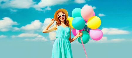 Happy smiling young woman with bunch of colorful balloons wearing a summer straw hat on the field on a blue sky backgroundの写真素材