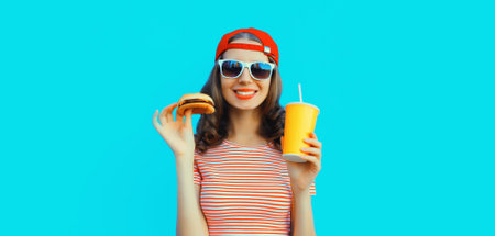 Portrait of stylish smiling young woman drinking juice with burger fast food on blue backgroundの写真素材