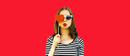 Portrait of beautiful young woman covering her eyes with red sweet heart shaped lollipop and blowing her lips on backgroundの写真素材