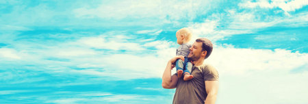 Happy strong father holding his son child on shoulder outdoors on blue sky background with clouds, blank copy space for advertising textの写真素材