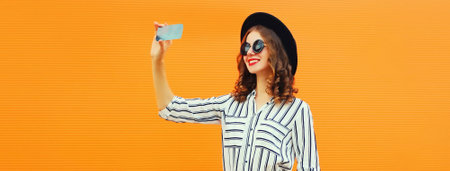 Portrait of happy smiling young woman taking selfie with smartphone wearing black round hat on orange backgroundの写真素材