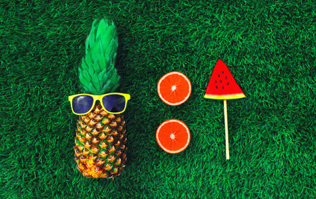 Summer fresh image of pineapple with sunglasses and lollipop or ice cream shaped slice of watermelon, orange or grapefruit on the grass backgroundの写真素材