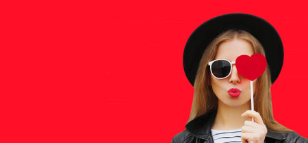 Portrait of stylish young woman with red heart shaped lollipop blowing her lips sending sweet air kiss wearing a black round hat on backgroundの写真素材
