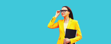 Portrait of young woman with folder wearing eyeglasses, yellow business suit on blue backgroundの写真素材