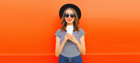 Portrait of happy smiling young woman drinking fresh juice or coffee wearing round black hat on orange backgroundの写真素材