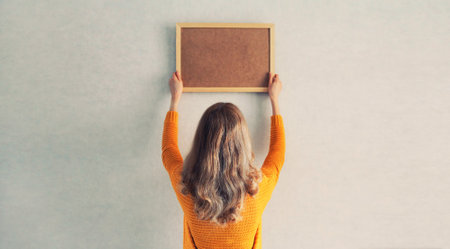 Young woman decorating interior, hanging blank a photo frame mockup on white wall in a new house
