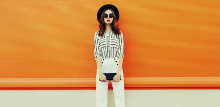 Portrait of beautiful young woman wearing white striped shirt, black round hat on orange background