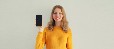 Portrait of happy smiling young woman showing black blank smartphone screen wearing knitted yellow...