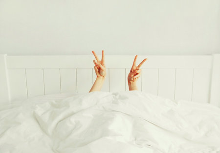 Cheerful lazy woman waking up after sleeping lying in soft comfortable bed showing gesture stretching her hands up from under the blanket in white bedroomの写真素材