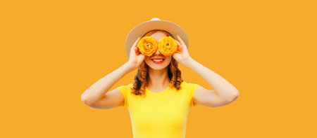 Summer portrait of happy smiling young woman covering her eyes with flower buds as binoculars looking for something wearing round straw hat on yellow backgroundの写真素材