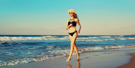 Summer vacation, happy smiling woman walking running in bikini swimsuit and straw hat on the beach on sea coast with waves background on sunny dayの写真素材
