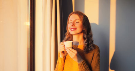 Portrait of happy satisfied smiling young woman enjoying hot tasty cup of coffee or tea in early...
