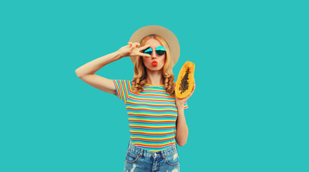 Summer portrait of young woman model posing with fresh papaya fruits wearing straw hat, sunglasses on blue backgroundの写真素材