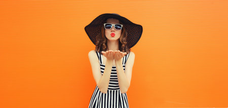 Summer portrait of beautiful young woman model blowing her lips sending sweet air kiss wearing black round straw hat on orange backgroundの写真素材