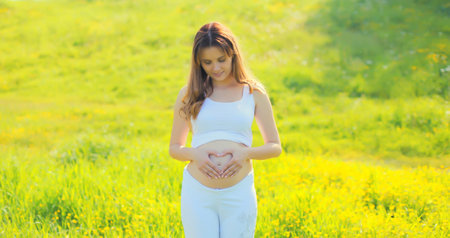 Happy young pregnant woman makes heart sign on her belly with her hands in summer park