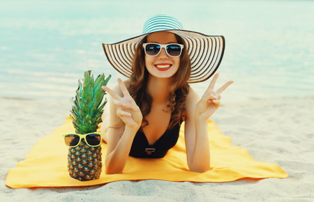 Summer vacation, happy young woman with pineapple fruits in bikini and straw hat lying on sand on the beach on sea backgroundの写真素材