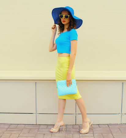 Beautiful young woman in full-length posing wearing summer straw hat, skirt and handbag clutch on city streetの写真素材