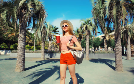 Beautiful young woman with cup of coffee in summer park wearing backpack, straw hat, shorts on palm tree backgroundの写真素材