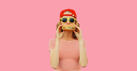Portrait of stylish smiling young woman with burger fast food on pink backgroundの写真素材
