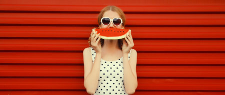 Summer portrait of happy smiling young woman eating fresh juicy fruits, slice of watermelon wearing heart shaped sunglasses on red backgroundの写真素材