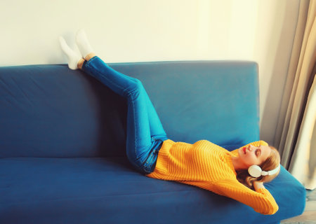Happy relaxed young woman listening to music with wireless headphones with closed eyes lies on the couch at homeの写真素材