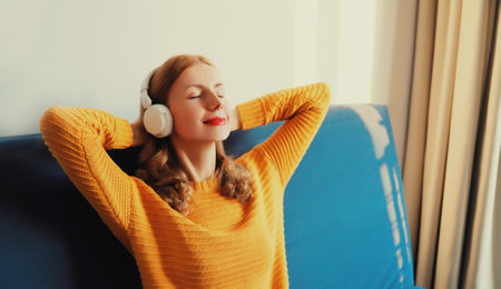 Happy relaxed young woman listening to music with wireless headphones with closed eyes lies on the couch at homeの写真素材