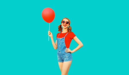 Happy cheerful young woman having fun with balloon wearing red heart shaped sunglasses, jumpsuit on blue backgroundの写真素材