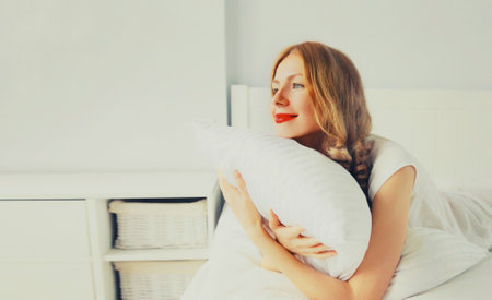 Healthy sleep, happy relaxed young woman hugs her pillow during lying on soft comfortable bed in white bedroomの写真素材