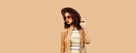 Portrait of beautiful young woman wearing jacket, sunglasses, round hat on brown wall studio backgroundの写真素材