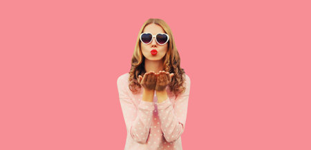 Portrait of beautiful young woman model blowing her lips with red lipstick sends sweet air kiss wearing white heart shaped sunglasses on pink backgroundの写真素材