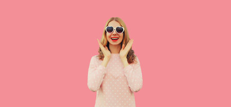 Portrait of happy excited surprised young woman wearing white heart shaped sunglasses on pink backgroundの写真素材