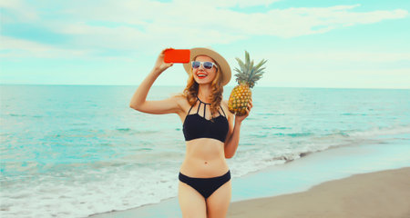 Summer vacation, happy smiling young woman holding pineapple taking selfie with smartphone wearing bikini swimsuit and straw hat on the beach on sea coast backgroundの写真素材
