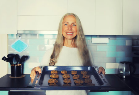 Portrait of happy smiling middle-aged woman housewife holding baking tray with cookies in the kitchen at homeの写真素材