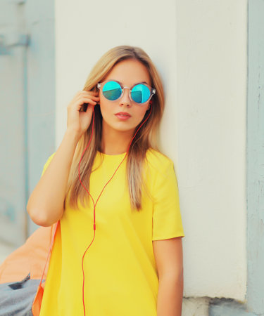 Portrait of stylish modern blonde young woman listening to music in headphones wearing sunglasses, backpack, yellow t-shirt in the cityの写真素材