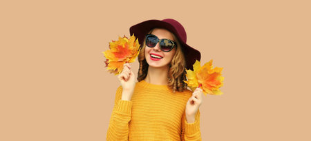 Autumn color style outfit, portrait of beautiful smiling young woman with yellow maple leaves wearing round hat, sweater on brown backgroundの写真素材